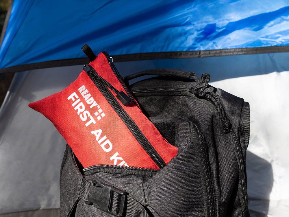 Red Ready First Aid kit in the pocket of a black backpack beside a tent